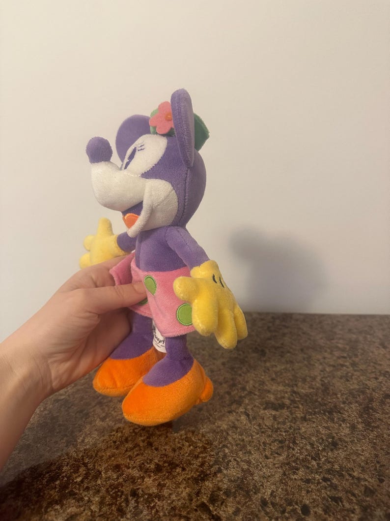 Pu&ograve; includere: Bambola di peluche di Minnie Mouse in un abito tropicale. La bambola ha testa e corpo viola, viso bianco, guanti gialli, scarpe arancioni e gonna rosa a pois verdi. Un fiore rosa e una foglia verde adornano la sua testa.