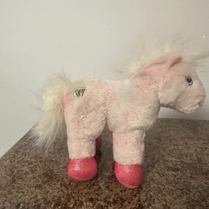 May include: A pink plush horse toy with a white mane and tail. The horse has pink glittery hooves and a small logo on its side. The toy is designed to look like a pony.