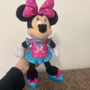 May include: A Minnie Mouse plush doll, featuring a pink dress with white polka dots, a matching bow, and blue accents. The doll has black ears, a black nose, and white gloves. The doll is approximately 10 inches tall.