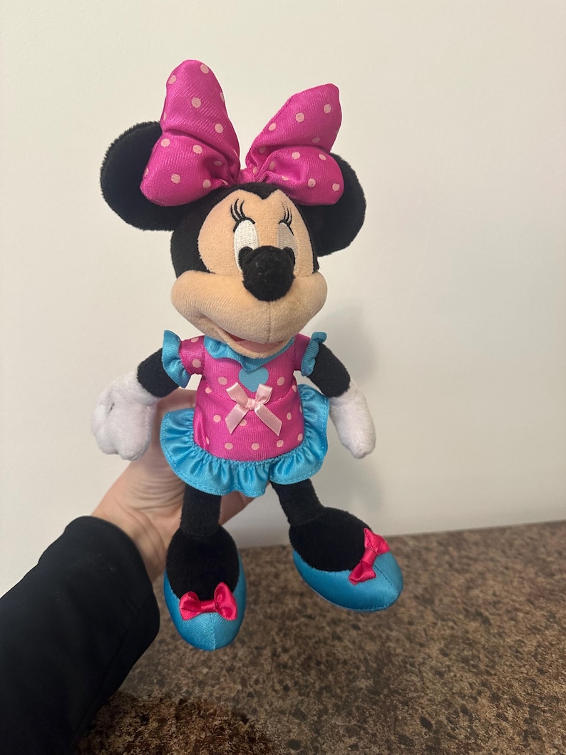 May include: A Minnie Mouse plush doll, featuring a pink dress with white polka dots, a matching bow, and blue accents. The doll has black ears, a black nose, and white gloves. The doll is approximately 10 inches tall.