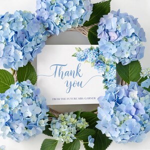 May include: A square white card with the blue text Thank you and FROM THE FUTURE MRS. GARNER, centered within a circular wreath of light blue hydrangea flowers and green leaves, featuring a matching floral accent on the top right corner.