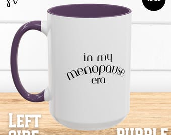May include: White ceramic mug with a purple interior and handle. The mug features the text "in my menopause era" in black script. The mug is 15 oz.