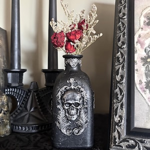 May include: Black decorative bottle with a skull design and dried red flowers. The bottle has a silver skull and leaf design. The bottle is part of a gothic home decor theme.
