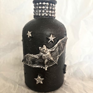 May include: Black decorative bottle with silver accents. The bottle features a silver bat and star embellishments. The top of the bottle is silver and textured.