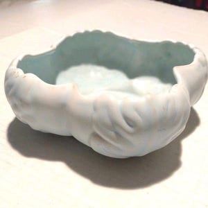 Puede incluir: Un cuenco de cerámica azul claro con forma de nube. El cuenco tiene un exterior liso y un interior azul claro. Los bordes son irregulares, lo que le da un diseño único. Está sobre una superficie blanca. Un objeto decorativo.
