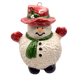 May include: A ceramic snowman ornament with a red hat, green scarf, and white body. The snowman has a carrot nose, black eyes, and a smiling mouth.