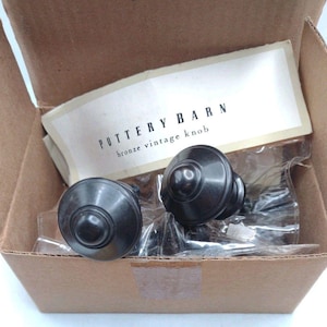 Puede incluir: Dos pomos vintage de bronce de Pottery Barn se muestran en una caja de cartón. Los pomos son redondos con un diseño central en relieve y están empaquetados en plástico transparente. El embalaje incluye una etiqueta que dice "POTTERY BARN bronze vintage knob."
