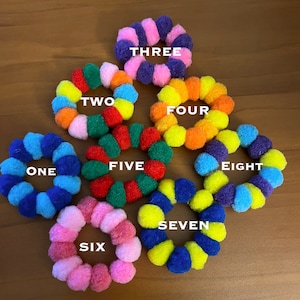 May include: Eight colorful pom-pom hair ties in a circle on a wooden surface. The hair ties are numbered one through eight.