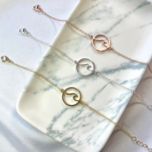 May include: Three wave bracelets on a white marble surface. The bracelets are made of silver, gold, and rose gold chains with a wave charm.
