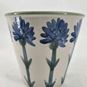 May include: A ceramic planter with a tapered cylindrical shape, featuring a cream-colored base and a hand-painted pattern of blue cornflowers with green stems and leaves. The rim of the planter is finished with a solid green painted band.