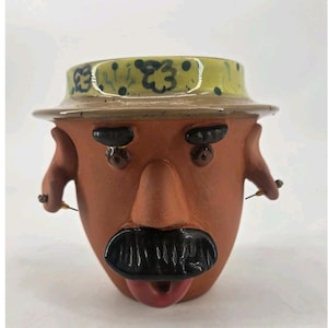 May include: A terracotta-colored ceramic planter shaped like a head, featuring a yellow hat with dark blue floral patterns, black eyebrows, a prominent nose, a thick black mustache, and a small red mouth. Two small metal earring posts are attached.