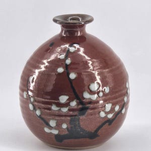 Pode incluir: Um vaso de cerâmica bordô com corpo redondo e pescoço estreito. O vaso é decorado com um galho preto e flores brancas. O vaso tem um acabamento brilhante.