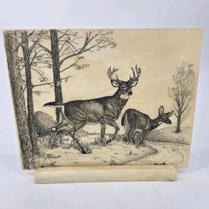 May include: A rectangular, off-white decorative plaque featuring a detailed black and white illustration of two deer in a forest setting. The larger deer has prominent antlers. The plaque is supported by a matching stand.