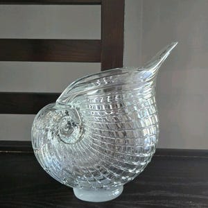 May include: A clear glass nautilus shell-shaped vase. The vase has a textured, ribbed design and a flared spout. The base is a solid, rounded shape. The vase is sitting on a dark surface, with a wooden chair in the background.