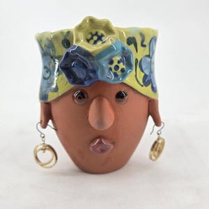 Może przedstawiać: Ceramiczna doniczka w kolorze terakoty w kształcie głowy, z żółtą opaską w niebieskie kwiatowe wzory i trójwymiarowymi akcentami kwiatowymi. Twarz ma wydatny nos, małe oczy i usta, a także złote kolczyki w kształcie kół.