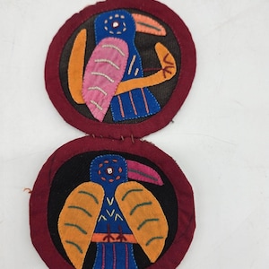 May include: Two circular fabric patches joined together, each featuring a blue bird with a pink or orange beak and yellow wings. The designs are stitched onto black fabric circles with dark red borders, showcasing detailed embroidery and applique.
