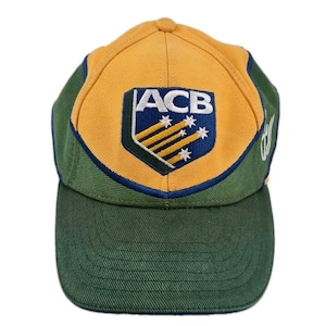 May include: A baseball cap with a green brim and a yellow top panel. The front features a shield-shaped emblem with the letters "ACB" and a design of stars and lines. The sides and back are green.