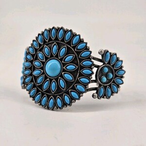 May include: Silver-toned cuff bracelet featuring a large, circular turquoise stone surrounded by smaller turquoise teardrop-shaped stones. The bracelet has an open design with a smaller floral accent on the side.