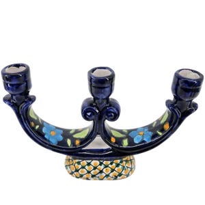 May include: A dark blue ceramic candelabra with three candle holders. The curved arms feature hand-painted light blue flowers with yellow centers and green leaves. The base has a yellow and green cross-hatched pattern on a white background.