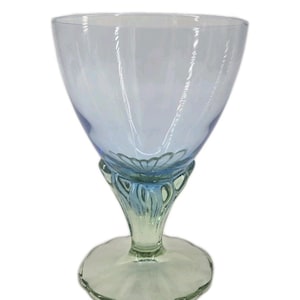 May include: A clear, stemmed glass with a light blue bowl and a decorative base. The base transitions from a light green to a blue hue, with a floral design at the stem's connection to the bowl. The glass is designed for beverages.