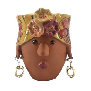 Pode incluir: Um vaso de cerâmica cor de terracota em forma de cabeça humana, com uma faixa amarela decorada com flores vermelhas pintadas. O rosto tem um nariz proeminente, olhos pequenos e uma boca rosa. Dois brincos de argola dourados pendem das orelhas.