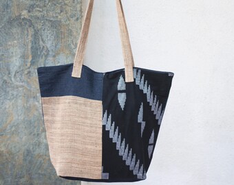 May include: A large tote bag with a patchwork design. The bag features a combination of denim blue, beige, and black fabric with a geometric pattern. It has long beige straps for carrying.