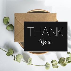 May include: A black card with white text that says "THANK YOU" in a modern font. The card is sitting on a brown envelope and a light brown plate.