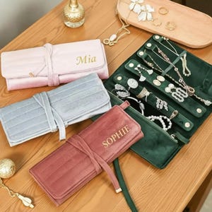 Puede incluir: Organizadores de joyas en rosa, azul y burdeos, con nombres personalizados. Un estuche enrollable de terciopelo verde abierto, mostrando collares y anillos. Los organizadores están sobre una superficie de madera.