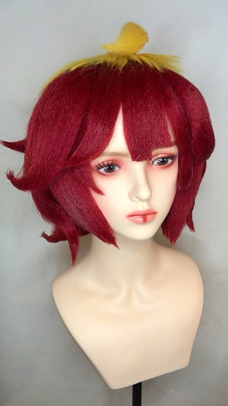 May include: A red wig with a yellow topknot. The wig is styled in a short bob with bangs. The wig is on a mannequin head.