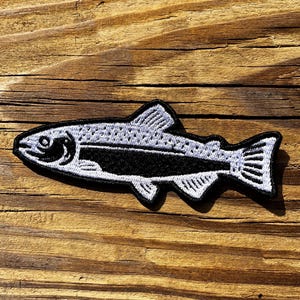 May include: A black and white embroidered patch in the shape of a fish. The fish has detailed stitching, including scales and fins. The patch is likely designed to be ironed or sewn onto fabric.