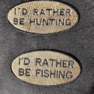 Peut inclure: Deux écussons brodés de forme ovale avec du fil doré. Le patch supérieur indique "I'D RATHER BE HUNTING" en texte noir. Le patch inférieur indique "I'D RATHER BE FISHING" en texte noir. Le fond est un tissu foncé et texturé.