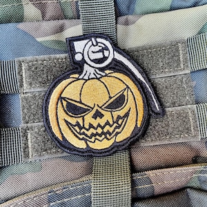 Puede incluir: Un parche temático de Halloween con un diseño de granada de calabaza. El parche tiene una calabaza amarilla con una cara tallada y un contorno negro. El fondo es un patrón de camuflaje.