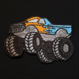 May include: Embroidered patch featuring a monster truck design. The truck has a gray body, orange and blue accents, and large black tires. The patch is detailed with stitching and is likely for application on clothing or accessories.