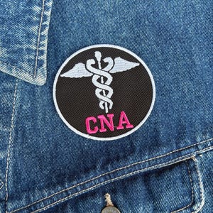 May include: A round patch with a black background, white border, and a white caduceus symbol with wings. The letters "CNA" are embroidered in pink. The patch is affixed to a blue denim jacket.