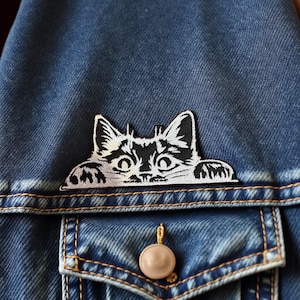 May include: A black and white cat patch is sewn onto a blue denim jacket. The patch features a stylized cat face with large eyes and paws. The jacket has a buttoned pocket and a beige button.