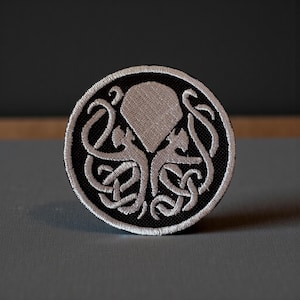 Może przedstawiać: Okrągła naszywka z haftem, czarne tło i biały, stylizowany wzór stworzenia głowonoga. Wzór zawiera skomplikowane sploty i białą obwódkę. Do odzieży lub akcesoriów.