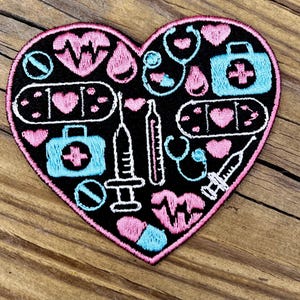 以下が含まれることがあります： 黒い背景とピンクの縁取りのハート型のパッチ。パッチには、心電図モニター、聴診器、救急箱、注射器など、ピンク、白、水色の医療をテーマにしたイラストが描かれています。パッチはおそらく刺繍されたアイテムです。