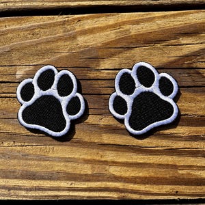 May include: Two embroidered paw print patches. Each patch features a black paw print with a white outline. The patches are displayed on a wooden surface with a natural wood grain.