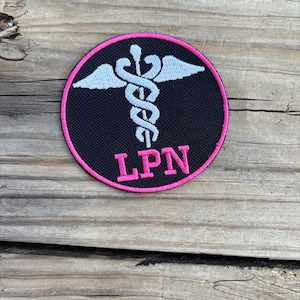 Può includere: Una toppa rotonda nera con un bordo rosa. La toppa presenta un simbolo di caduceo bianco e le lettere rosa "LPN" in basso. Lo sfondo è una superficie di legno.
