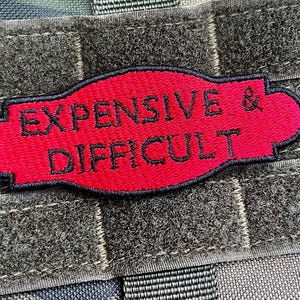 Op de afbeelding: Een rood geborduurd embleem met de zwarte woorden "EXPENSIVE & DIFFICULT". De patch heeft een zwarte rand en is geplaatst tegen een grijze, getextureerde achtergrond met klittenbandriemen.