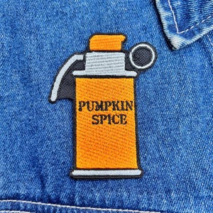 Puede incluir: Parche bordado con forma de granada, con cuerpo naranja y detalles grises y negros. El texto "PUMPKIN SPICE" está bordado en negro sobre el cuerpo naranja. El parche está adherido a una chaqueta vaquera azul.