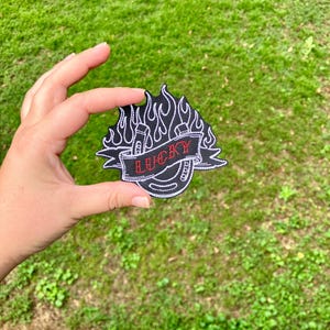 May include: Black and white embroidered patch with a horseshoe, flames, and the word "LUCKY" in red.