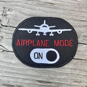 May include: Black oval patch with the words "AIRPLANE MODE" in red and "ON" in white. A white airplane graphic is above the text. The patch is on a wooden surface.