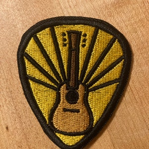 Może przedstawiać: Naszywka haftowana na ż&oacute;łto i czarno w kształcie kostki do gitary z brązowym akustycznym gitarą w środku. Gitara jest otoczona ż&oacute;łtymi promieniami światła.