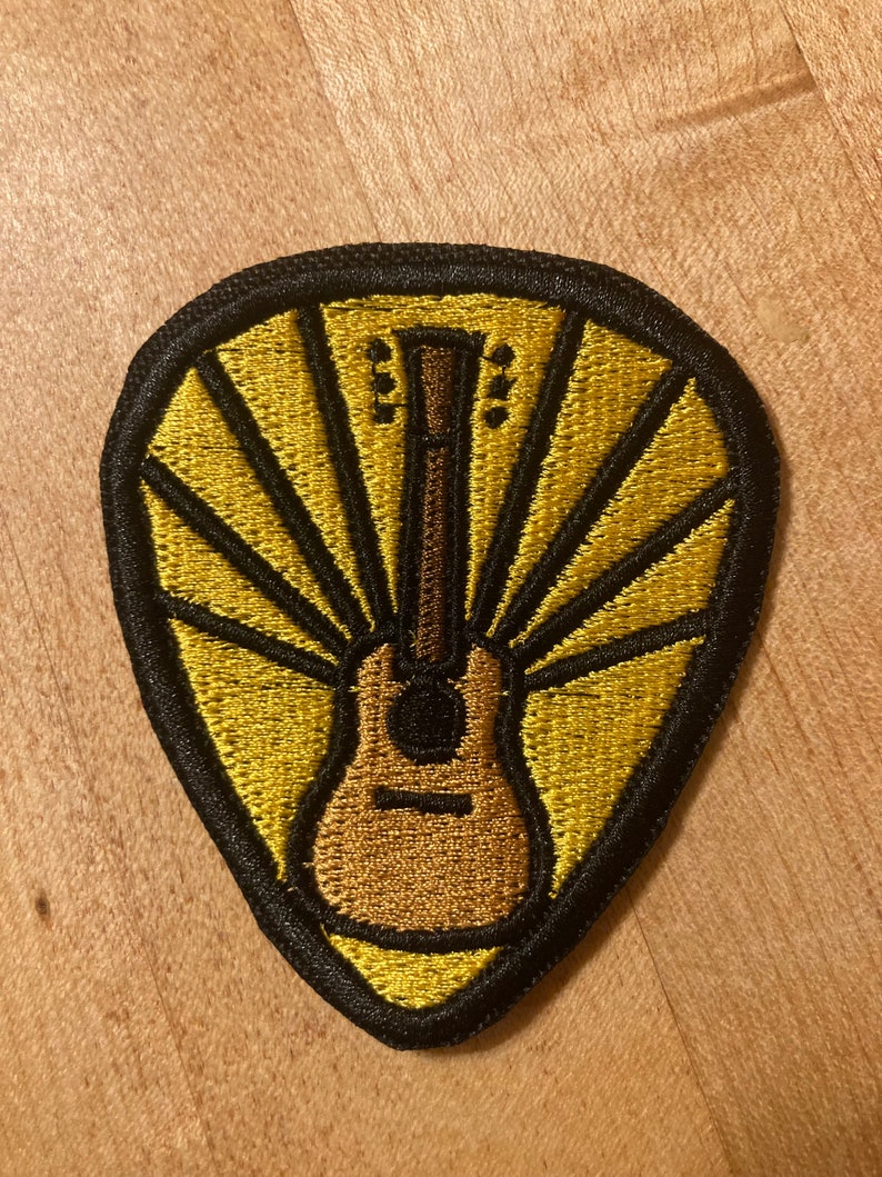 Może przedstawiać: Naszywka haftowana na ż&oacute;łto i czarno w kształcie kostki do gitary z brązowym akustycznym gitarą w środku. Gitara jest otoczona ż&oacute;łtymi promieniami światła.
