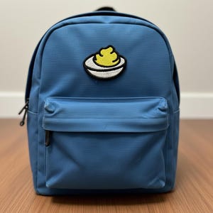 Puede incluir: Una mochila azul con un bolsillo frontal y un asa superior. La mochila presenta un parche de un alimento amarillo sobre un plato blanco con un contorno negro. La mochila tiene cremalleras negras.