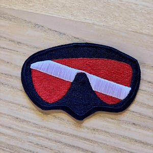 May include: A black, red, and white scuba diving mask patch. The patch is made of fabric and has a stitched design.