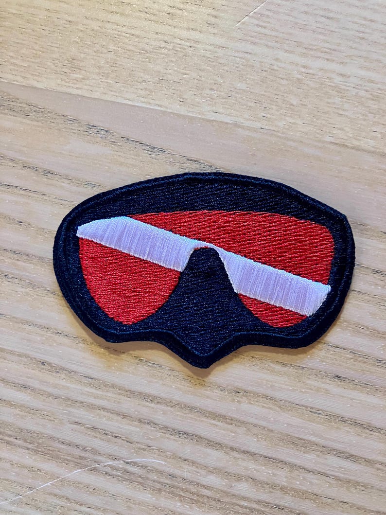 May include: A black, red, and white scuba diving mask patch. The patch is made of fabric and has a stitched design.