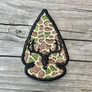 May include: Embroidered patch in the shape of an arrowhead, featuring a deer skull and antlers. The patch has a camouflage pattern in shades of brown, green, and tan, outlined with black stitching. The deer skull and antlers are black.