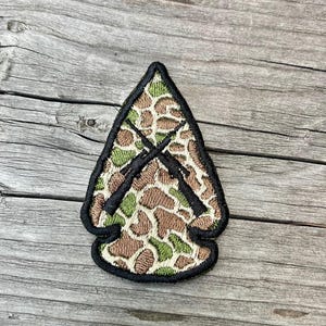 May include: Arrowhead-shaped patch with a camouflage pattern in shades of brown and green. Two black rifles are crossed in the center. The patch has a black border and is set against a wooden background.